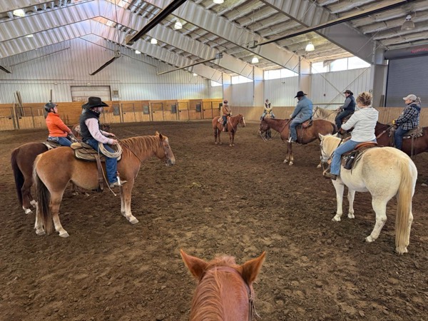 Seen between a horse’s ears, riders sit in a circle in an indoor arena